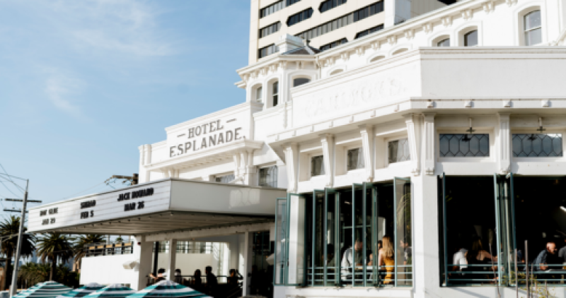 The exterior of the Hotel Esplanade building, with blue sky in the background and people seated at tables inside the open windows in the afternoon sun.